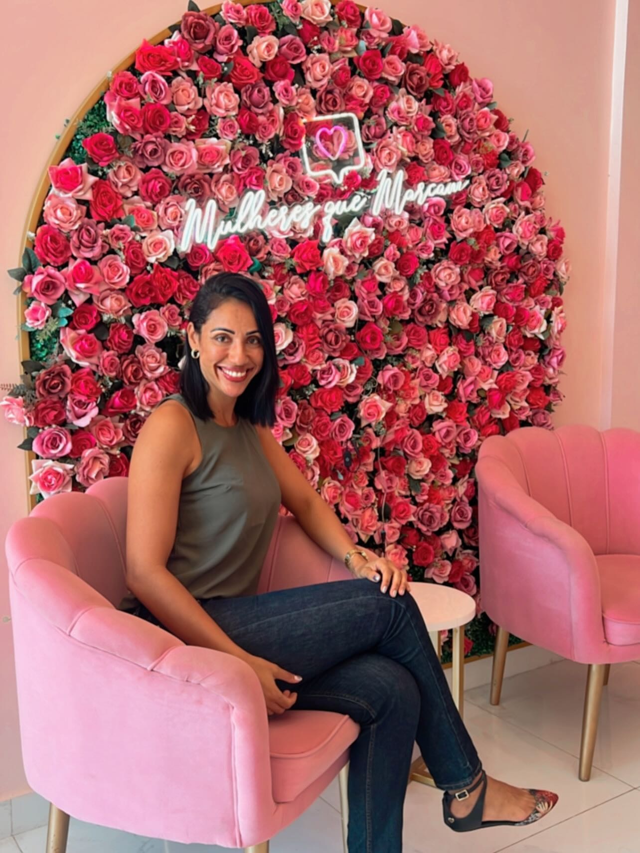Dra. Jaqueline Martins sentada na poltrona rosa do consultório, com parede de flores e letreiro de neon ao fundo